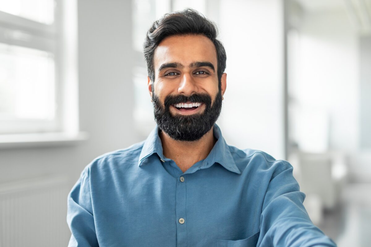 Cheerful smiling indian man wearing shirt sitting at office, closeup photo of happy businessman at